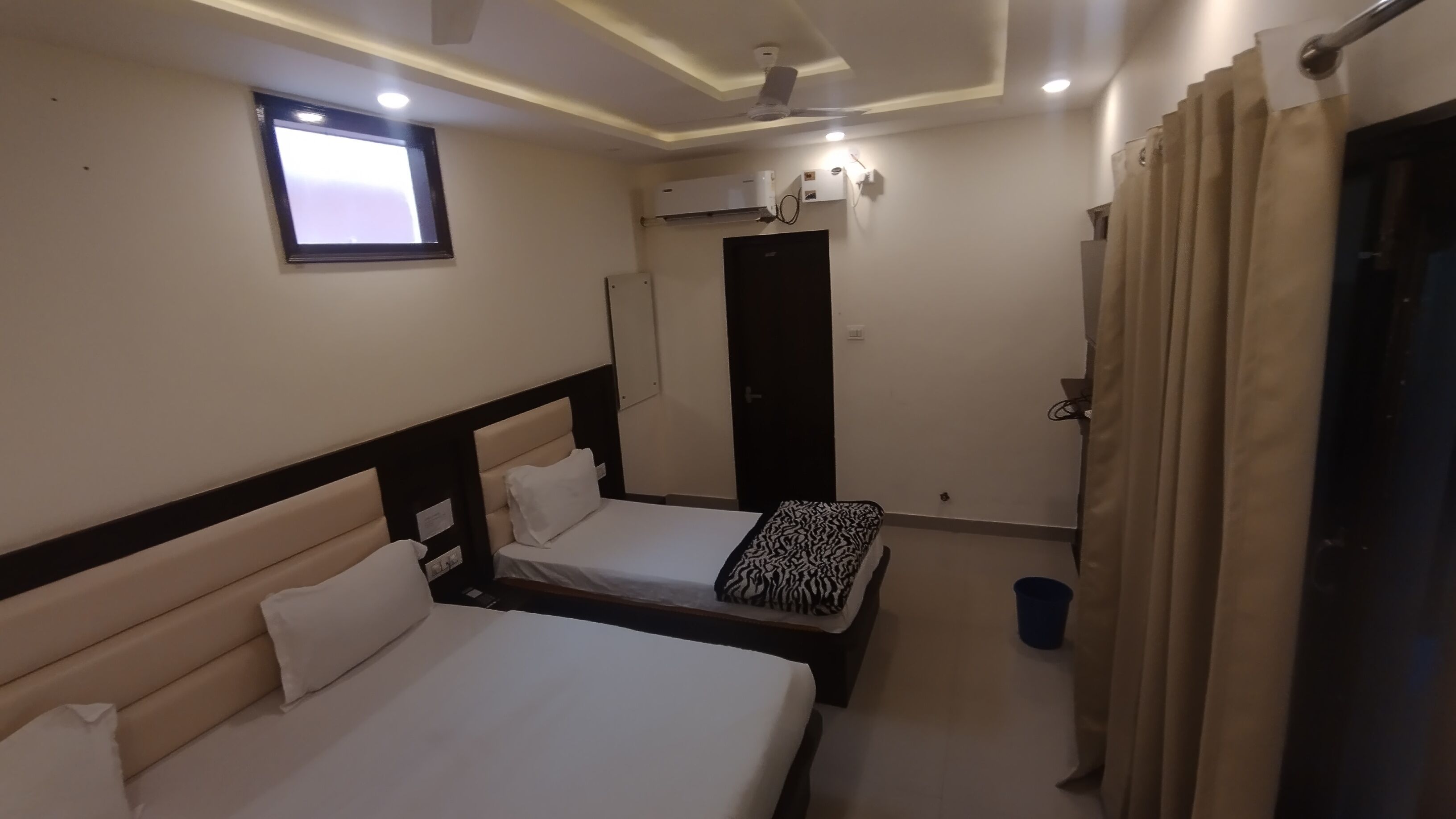 A hotel room with two beds, a window, an air conditioning unit, and a door.