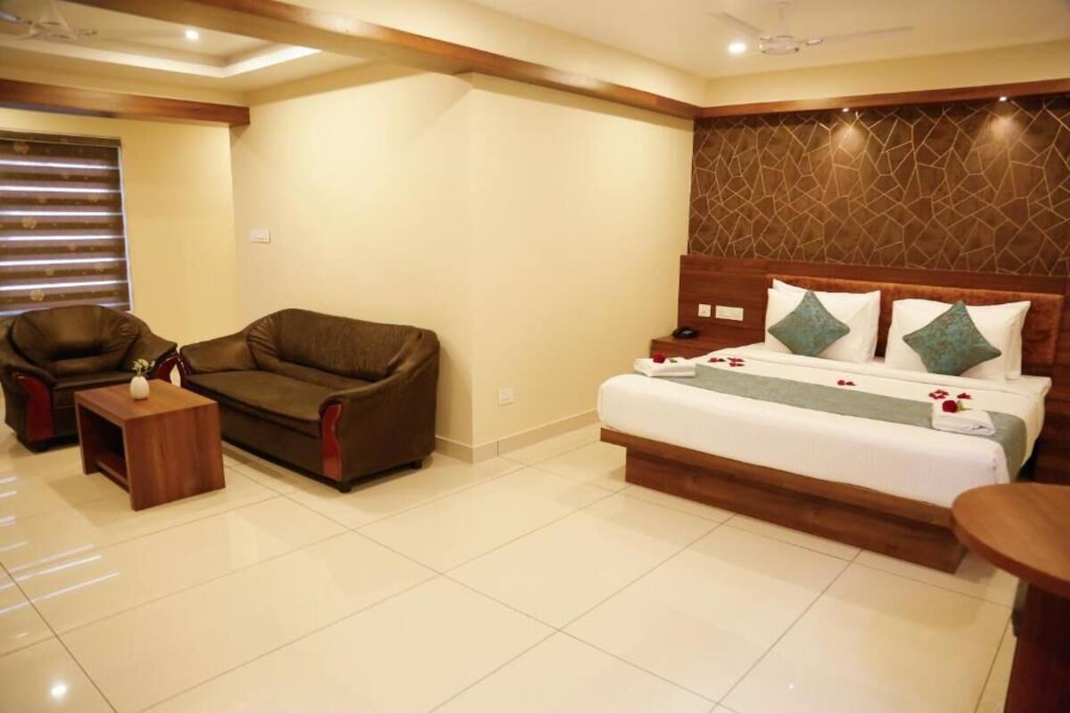 A hotel room with a large bed, a sofa, a small table, and a window with blinds.