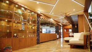 Um hall de hotel moderno com uma vitrine de vidro cheia de objetos decorativos, uma escadaria para o andar superior e uma área de estar com sofá e poltronas.