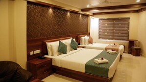 Quarto de hotel com duas camas, cabeceiras de madeira, uma janela grande com persianas, uma mesa de cabeceira com abajur e um pequeno vaso de planta.