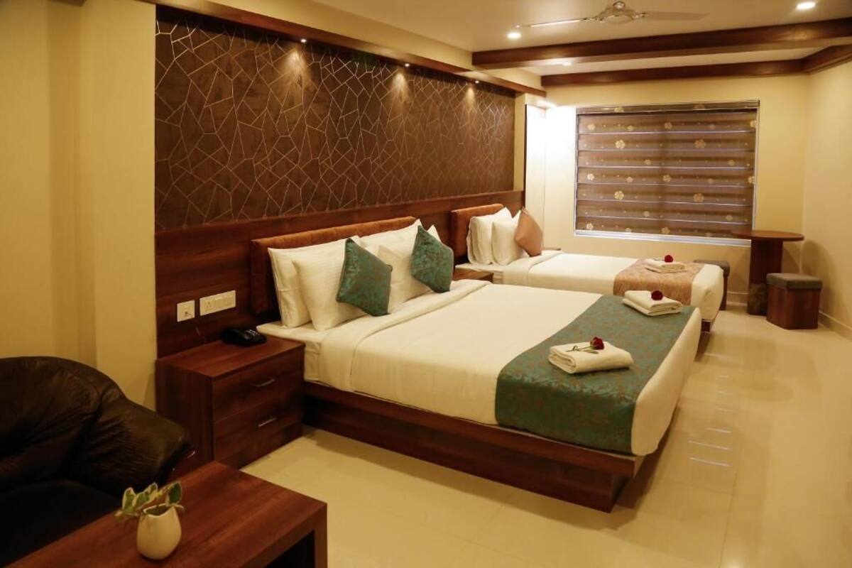 A hotel room with two beds, wooden headboards, a large window with blinds, a bedside table with a lamp, and a small potted plant.