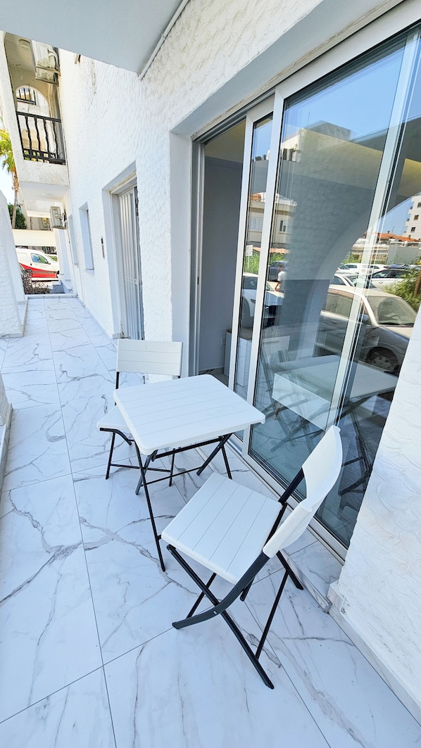 An outdoor balcony with a white table and two chairs, a sliding glass door, and a view of parked cars and buildings.