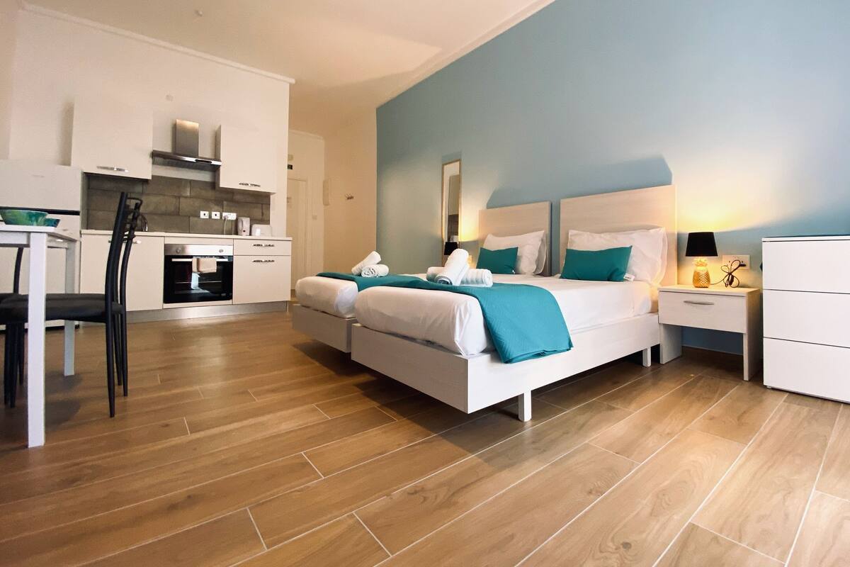 A modern hotel room with a kitchenette, a bed with teal bedding, and a nightstand with a lamp.