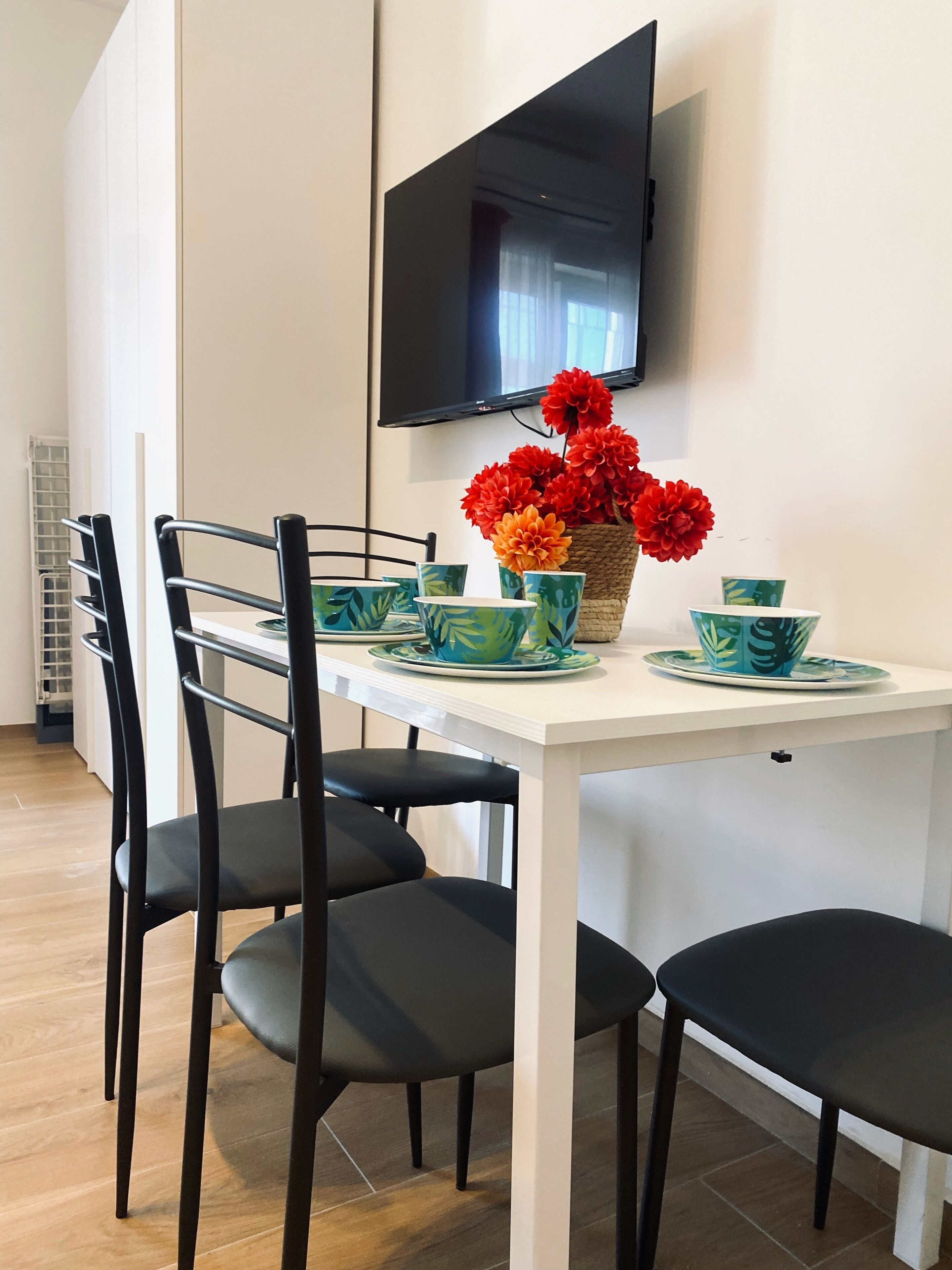 A dining area with a white table, black chairs, green and blue patterned tableware, and a basket of red flowers.