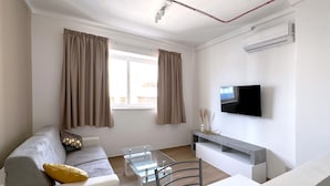 A modern living room with a sofa, a glass coffee table, a TV mounted on the wall, and a window with curtains. - Miami Apartments (St. Julian's)