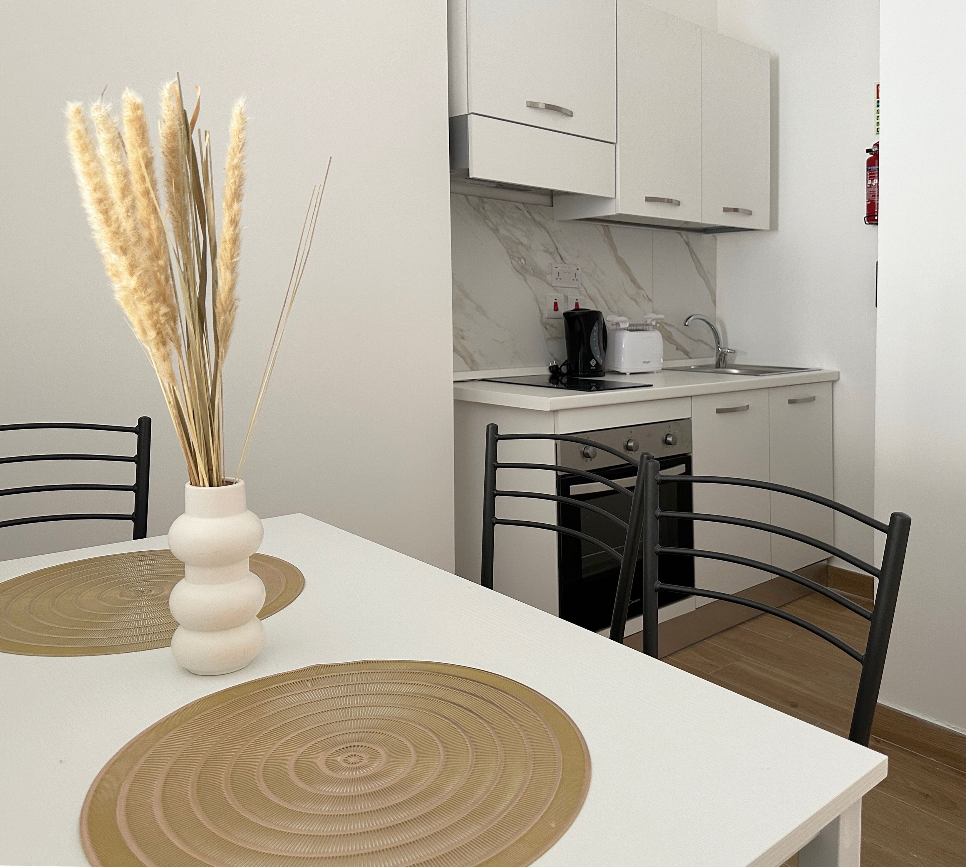 A modern kitchen with white cabinets, a coffee maker, and a toaster. There is a dining table with two chairs and a vase with dried plants.