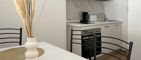 A modern kitchen with white cabinets, a coffee maker, and a toaster. There is a dining table with two chairs and a vase with dried plants.