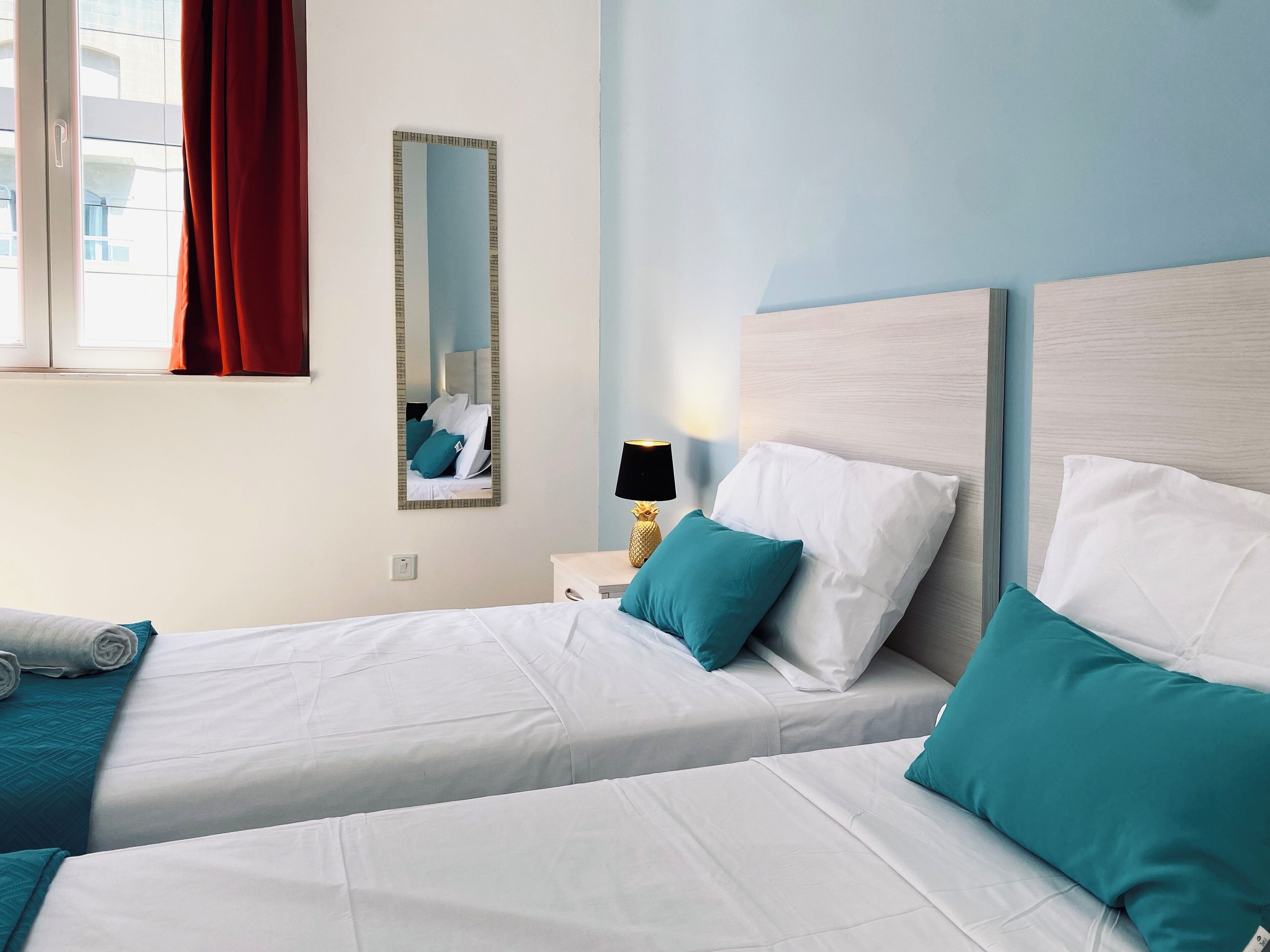 Una camera d'albergo con due letti, un comodino con una lampada, uno specchio e una finestra con tende.