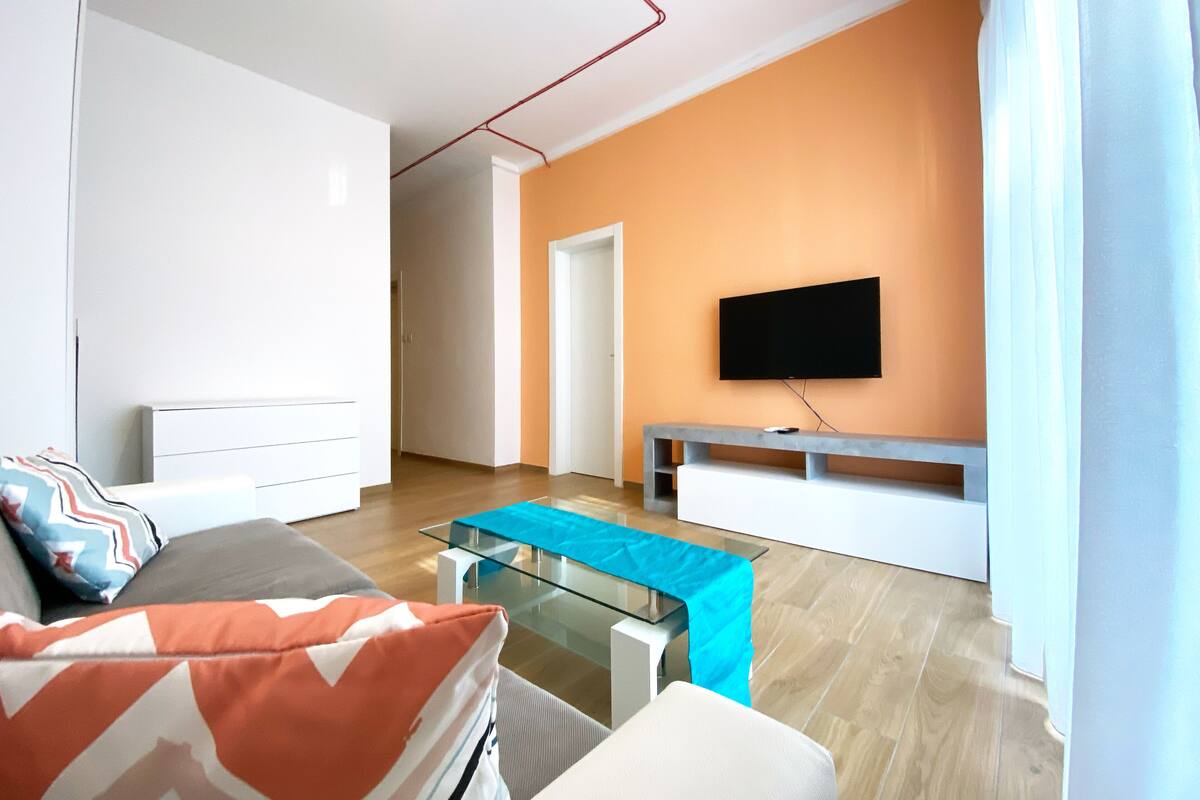A modern living room with a sofa, a glass coffee table, a TV mounted on the wall, and a wooden floor.