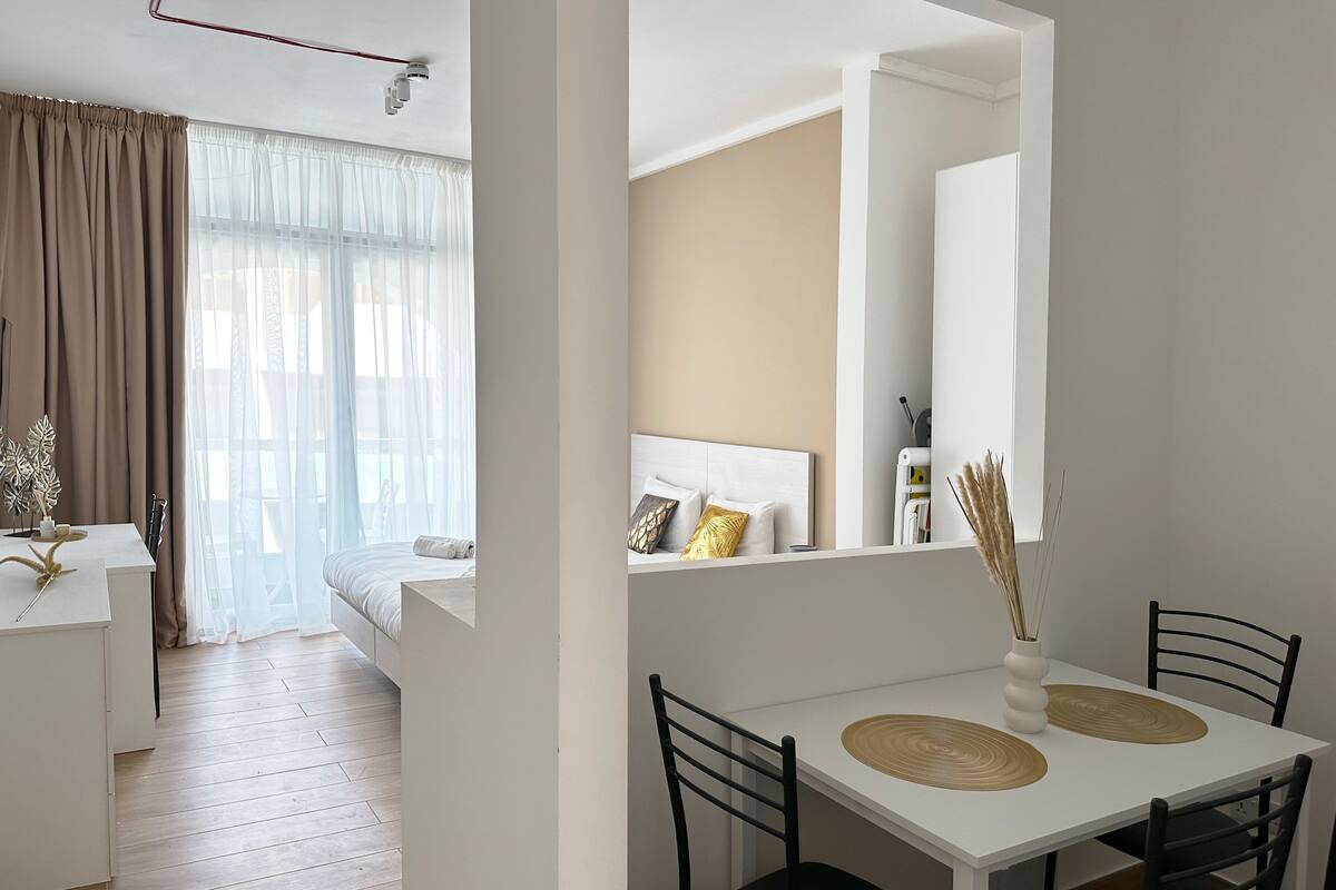 A modern hotel room with a bed, a small desk, a dining table with chairs, and a large mirror.