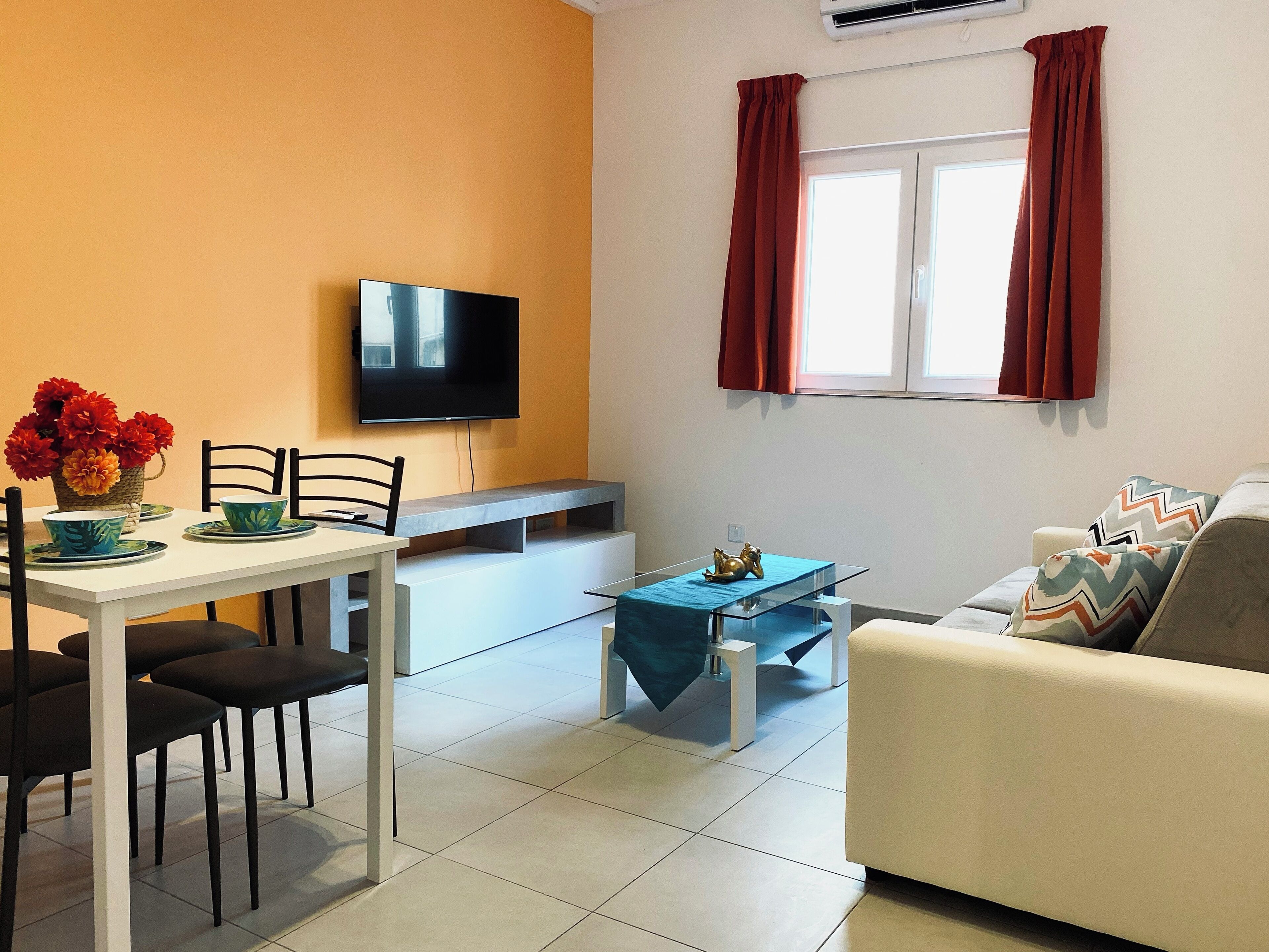 Uma sala de estar com televisão, uma mesa de jantar com cadeiras, um sofá e uma janela com cortinas.