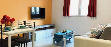 Uma sala de estar com televisão, uma mesa de jantar com cadeiras, um sofá e uma janela com cortinas.