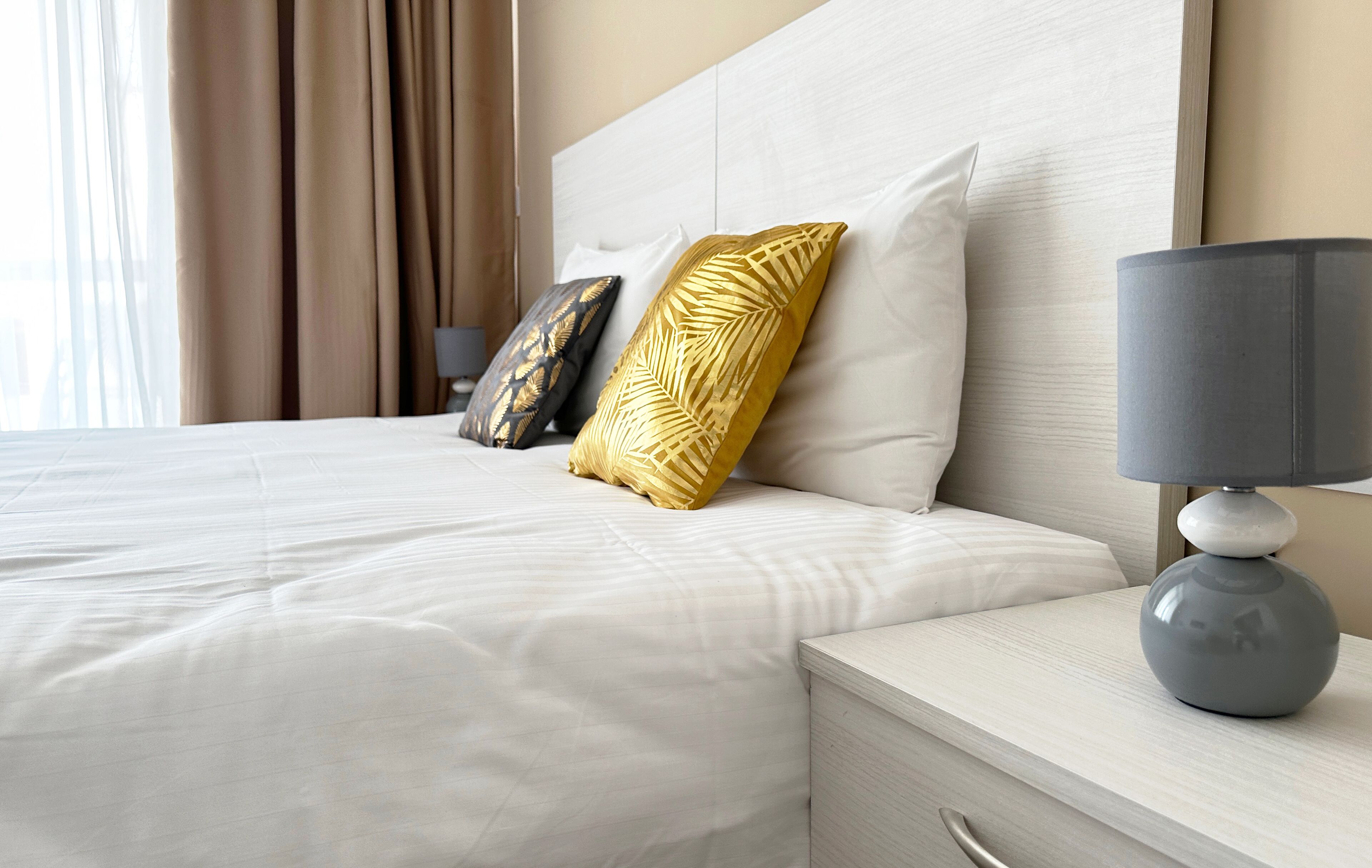 Una cama bien hecha con ropa blanca, dos almohadas decorativas, una mesita de noche con lámpara y vistas a la ventana con cortinas.