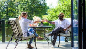 Deux personnes sont assises à une table, devant une bouteille et des verres, et elles ont une vue sur une étendue d’eau et sur la verdure environnante.