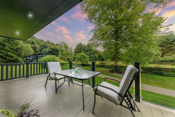 A covered patio with a table and chairs, overlooking a well-maintained garden and a body of water. - Secret Waterside Lodges in Worcestershire (Worcester)