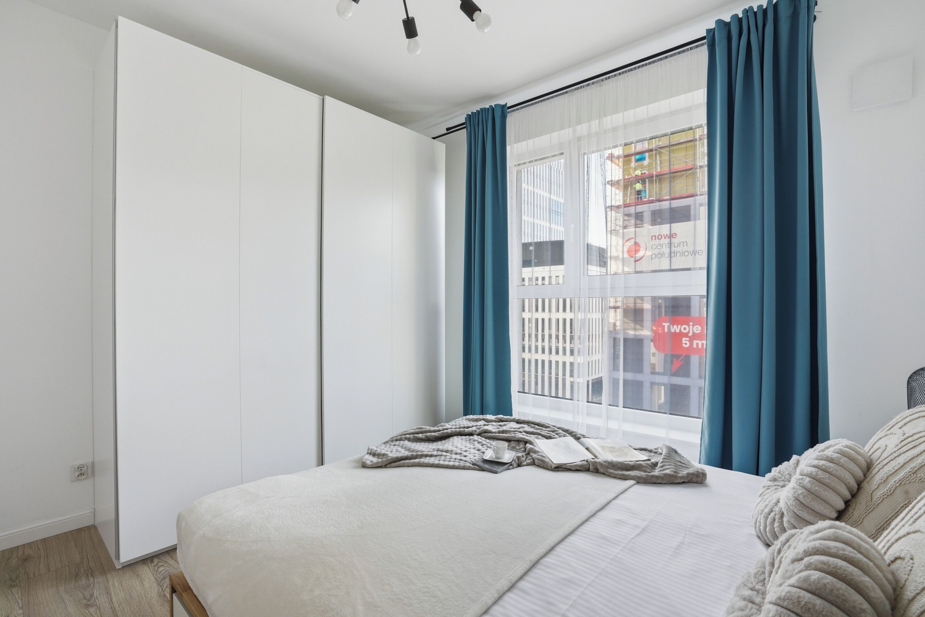 A modern bedroom with a large bed, a view of a building through sheer curtains, and a window with a view of a construction site.