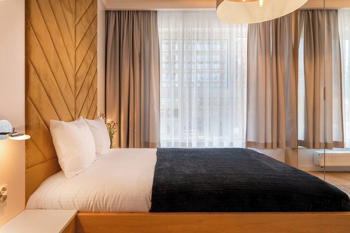 Habitación de hotel moderna con cabecera de madera, cama grande con ropa de cama blanca y negra, mesita de noche con lámpara y cortinas translúcidas que dejan entrar luz natural.