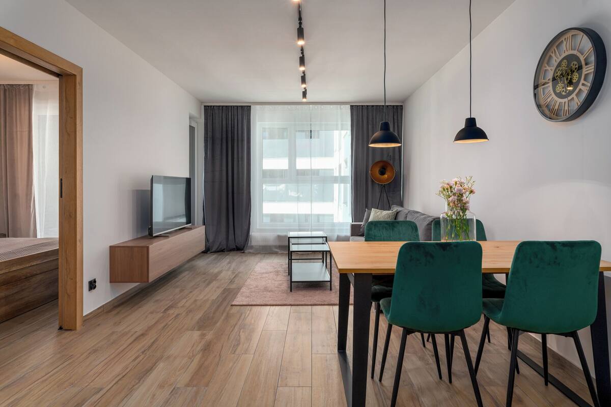 Una sala moderna con suelo de madera, televisor, mesa de comedor con sillas y un gran ventanal con cortinas.