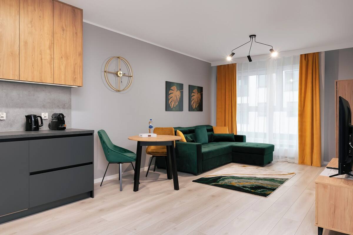 Un apartamento moderno con cocina, comedor y sala de estar. Cuenta con un mueble de madera, una mesa redonda con sillas, un sofá verde y un televisor sobre un mueble de madera.