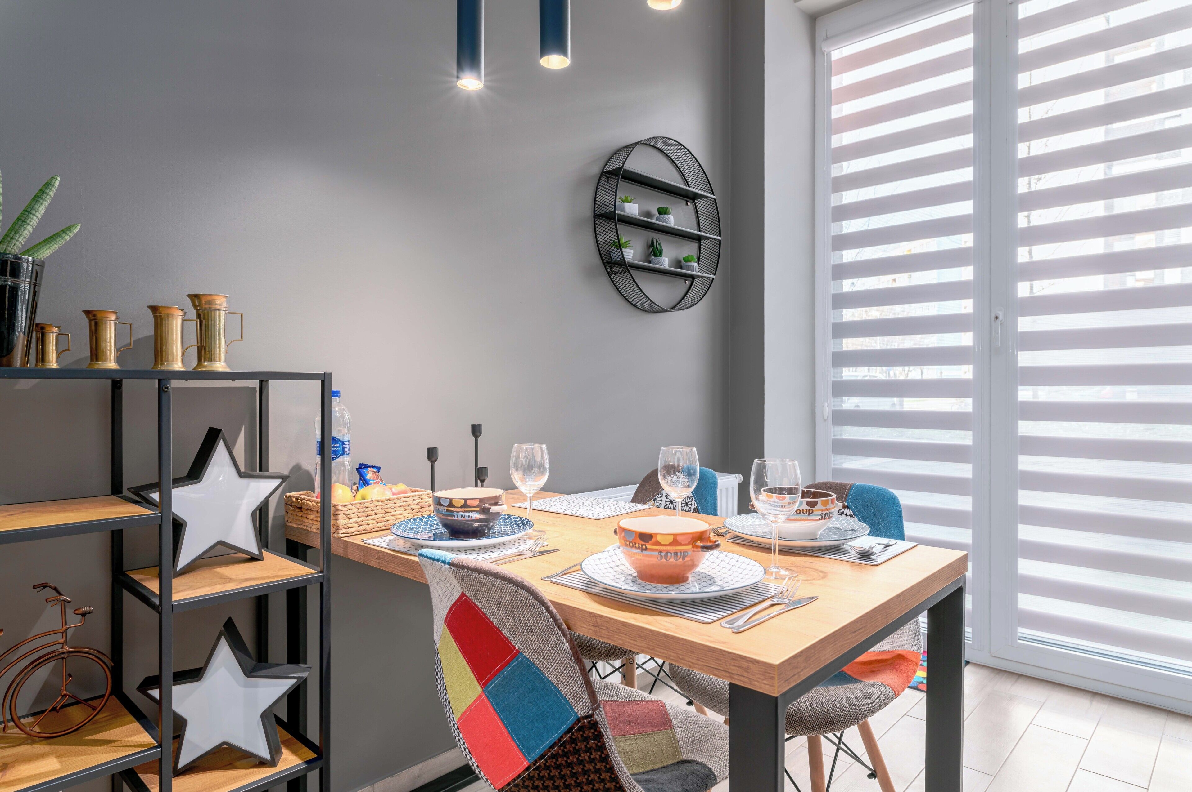 A dining area with a wooden table set for two, a black metal shelf with plants, and a window with blinds.