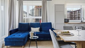 A modern living room with a blue sofa, a dining table with a plate of food and a glass, and a view of a cityscape through large windows. - Pure Rental Premium Apartments (Wroclaw)