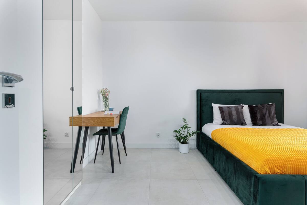 Un dormitorio moderno con una cama tapizada en verde, un escritorio de madera con una silla y una pequeña planta en maceta.