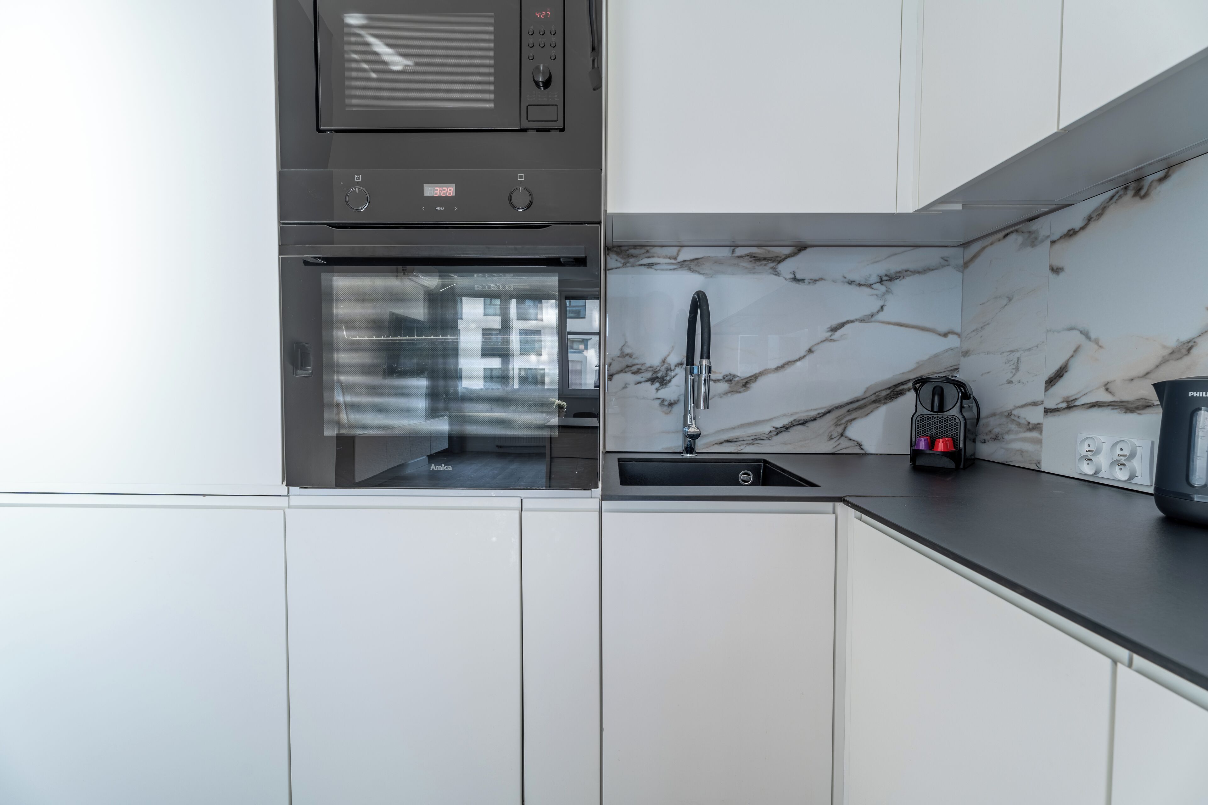 Een moderne keuken met witte kasten, een zwart aanrecht, een magnetron, een oven en een spoelbak met een marmeren achterwand.
