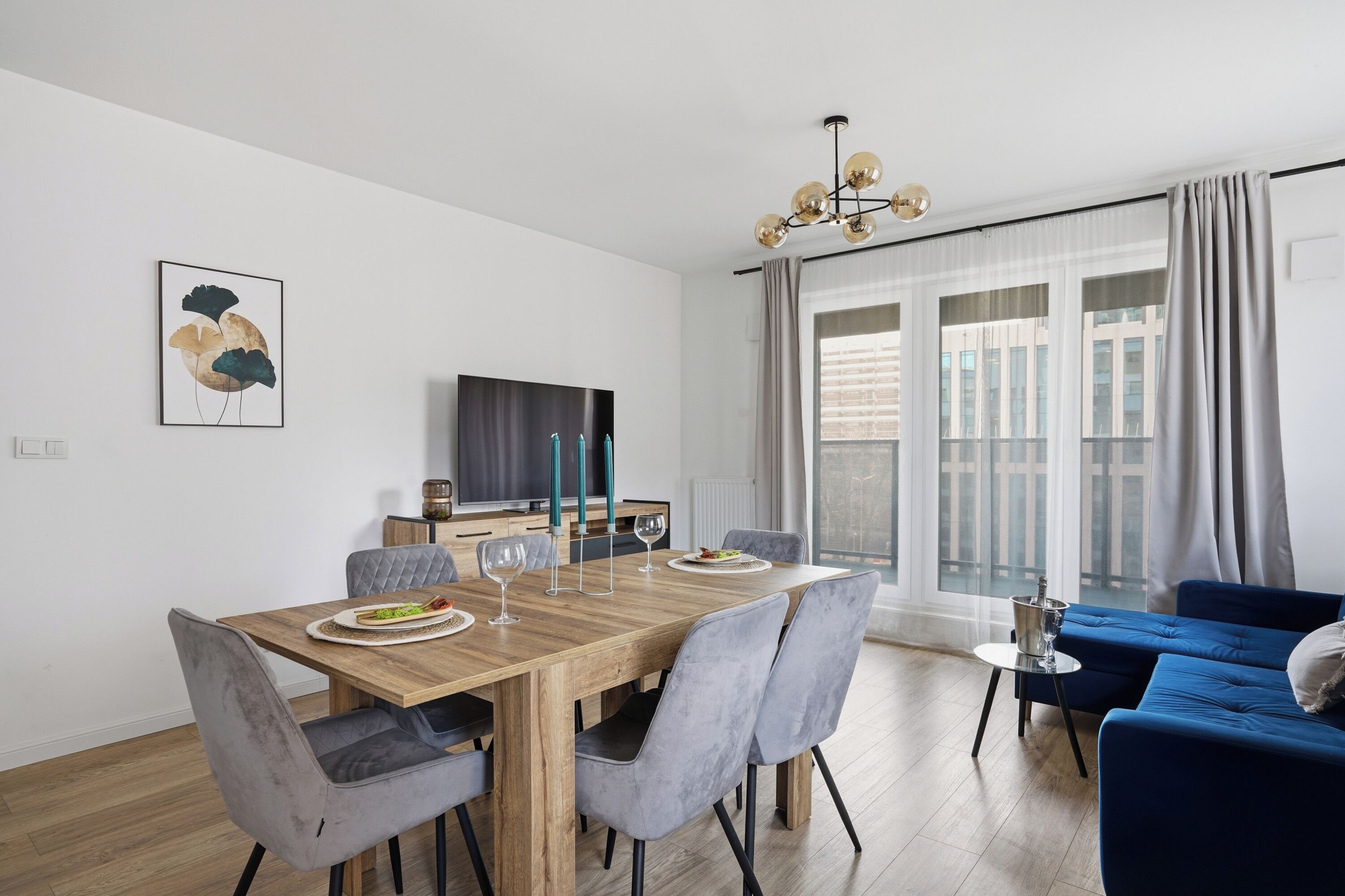 A modern living room with a wooden dining table, grey upholstered chairs, a blue sofa, and a large window with curtains.