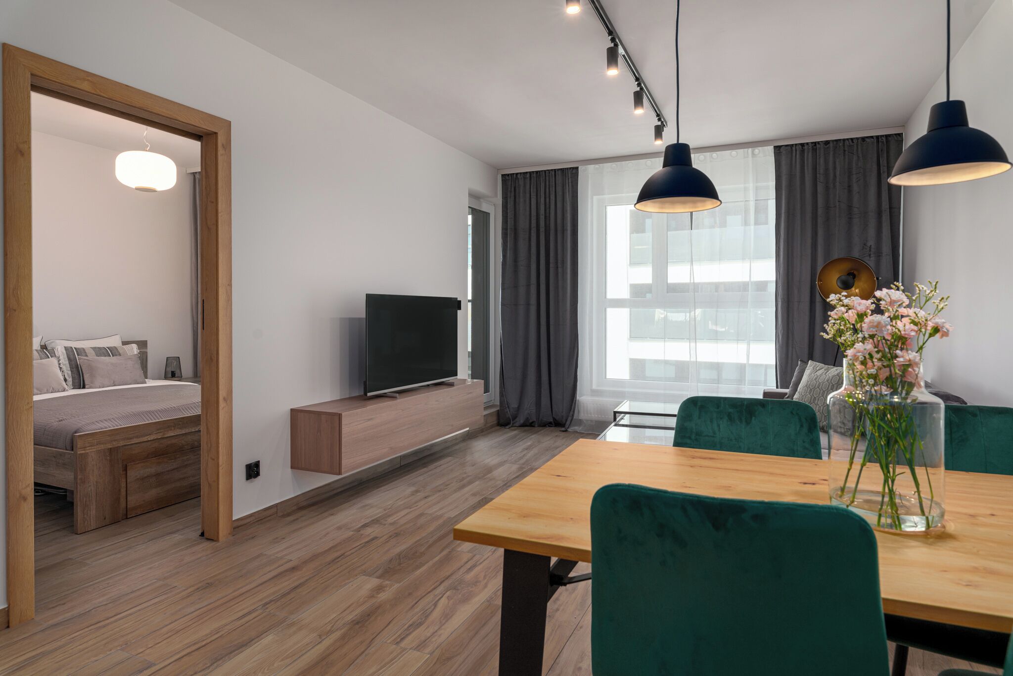 Een moderne woonkamer met een houten vloer, een eettafel met groene stoelen, een tv op een houten dressoir, en een groot raam met gordijnen.