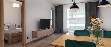 A modern living room with a wooden floor, a dining table with green chairs, a TV on a wooden stand, and a large window with curtains.