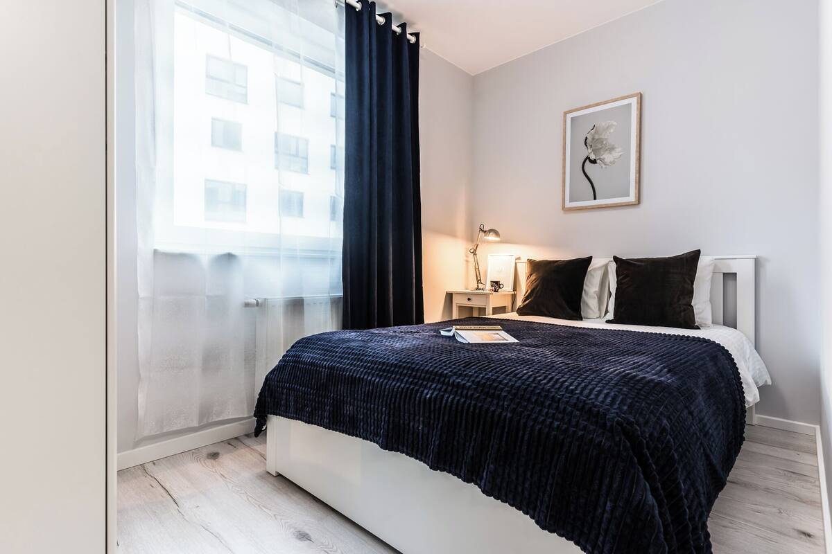 Un dormitorio con cama, mesita de noche, lámpara, un cuadro enmarcado en la pared y una ventana con cortinas.