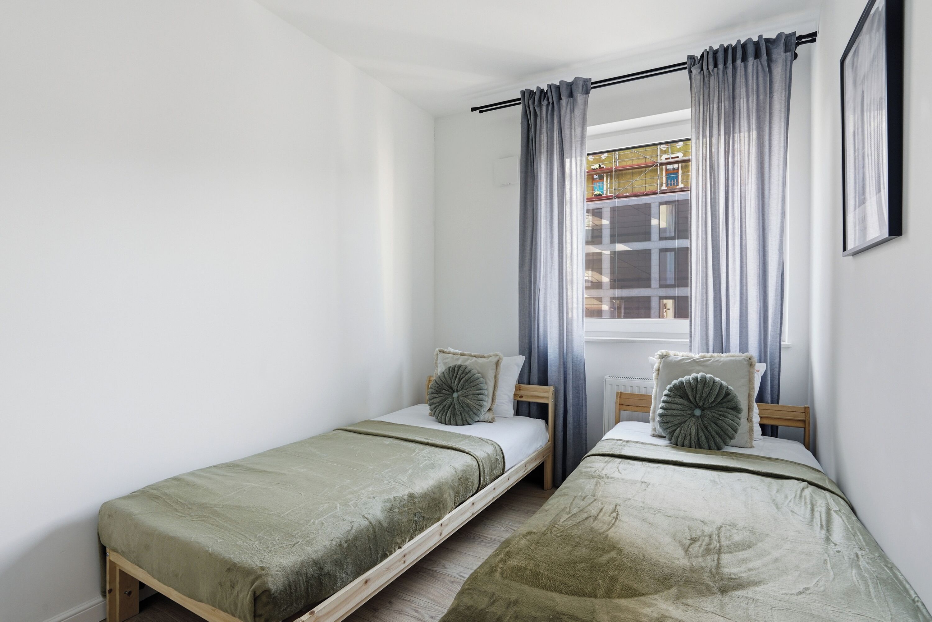 Une chambre avec deux lits simples, chacun doté d’un cadre en bois et de draps verts. Il y a une fenêtre avec des rideaux et un tableau encadré au mur.