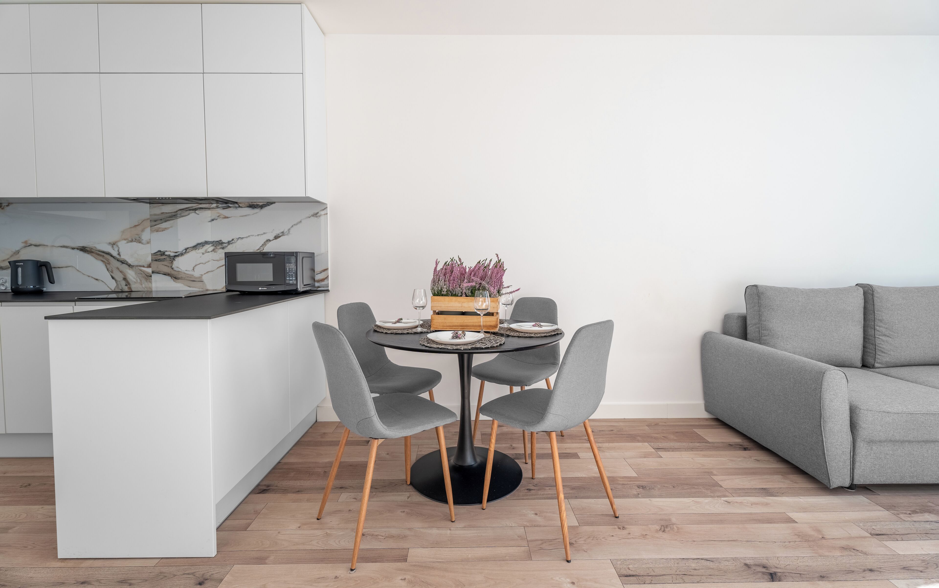 A modern kitchen with a built-in microwave, a dining table with four chairs, and a grey sofa in a living area.