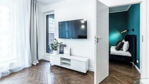 A modern living room with a flat-screen TV mounted on the wall, a white entertainment unit, a bed with dark blue bedding, and a teal accent wall. - Pure Rental Premium Apartments (Wroclaw)