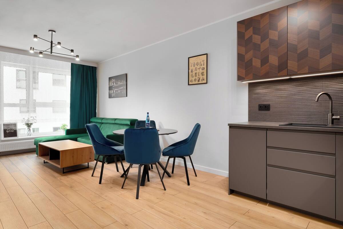 Una cocina moderna con estufa integrada, un lavamanos y un protector contra salpicaduras de madera. Hay una zona de comedor con un sofá verde y sillas azules.