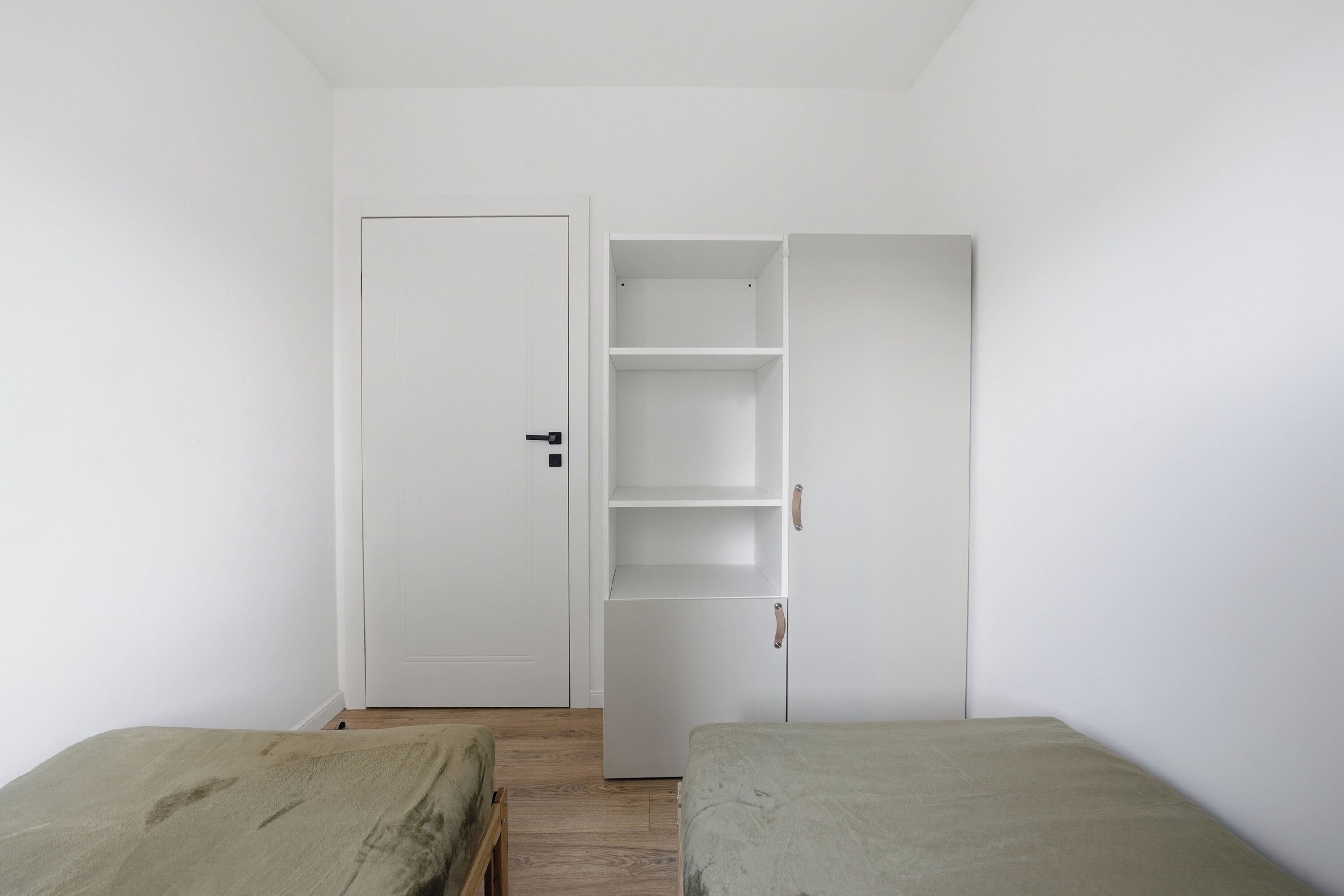 Una habitación con dos camas, un armario blanco con repisas abiertas y una puerta cerrada.