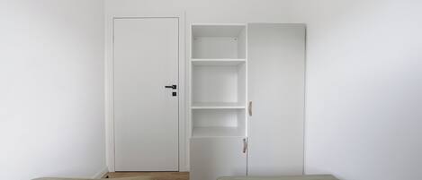 Una habitación con dos camas, un armario blanco con repisas abiertas y una puerta cerrada.