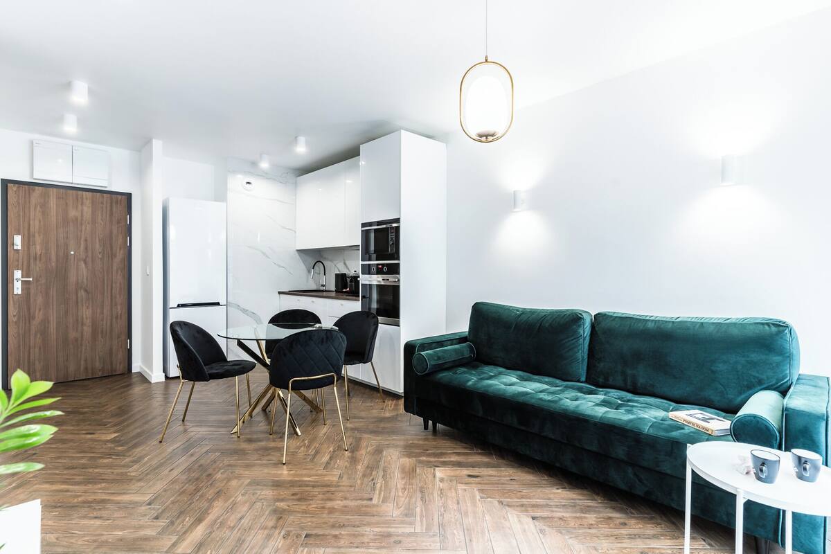 Una sala de estar moderna con un sofá verde, una mesa de comedor con sillas y una zona de cocina con armarios y electrodomésticos blancos.
