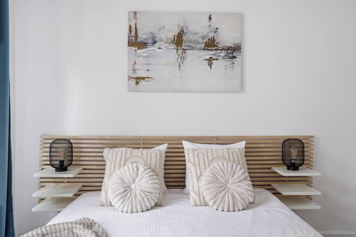 Un dormitorio con cabecera de madera de listones, dos mesitas de noche con lámparas negras, y un cuadro abstracto grande en la pared.