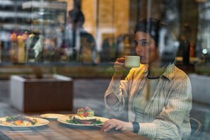 A person sitting at a table with a plate of food and a cup, in a cafe with a glass window. - Pure Rental Premium Apartments (Wroclaw)