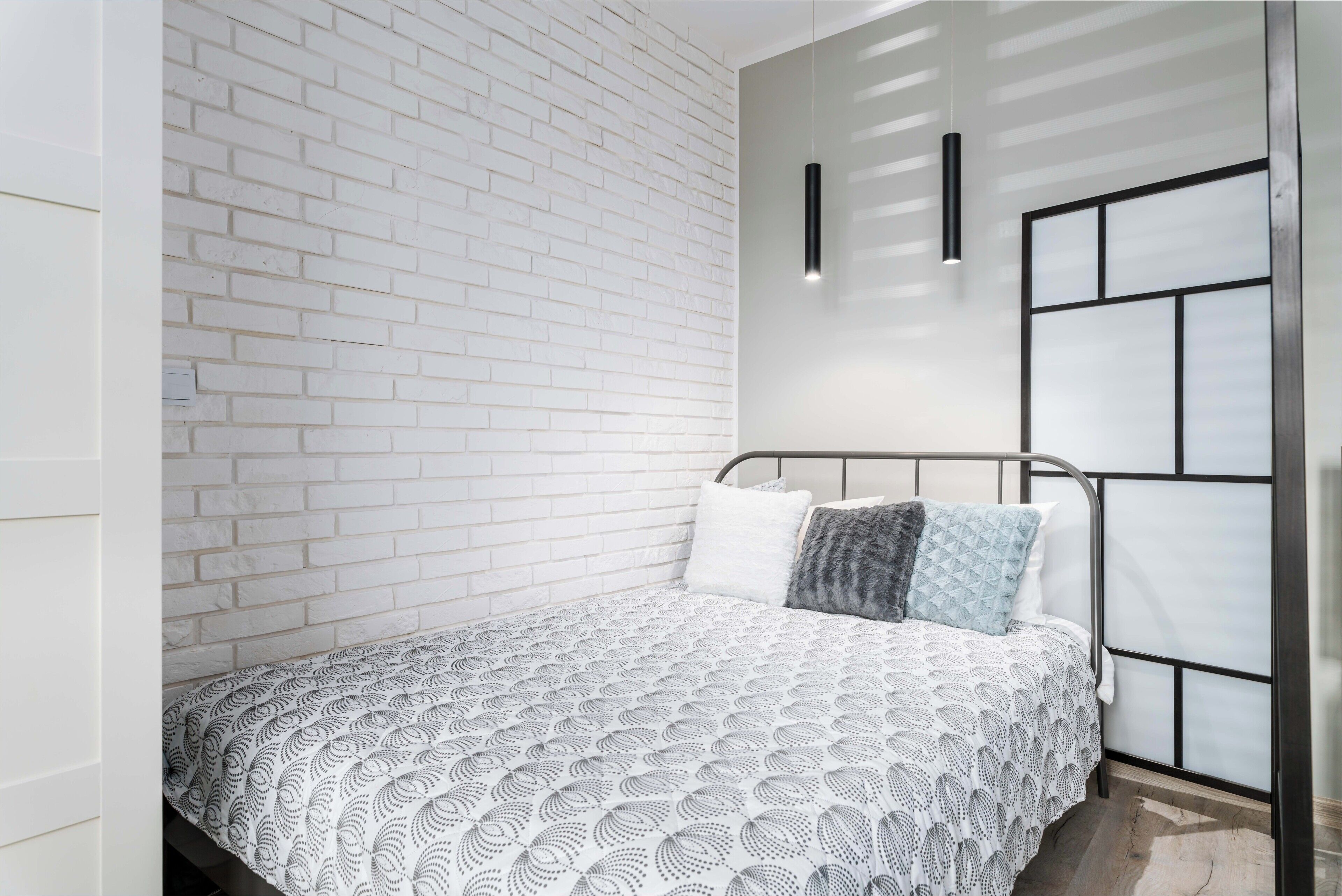 A bedroom with a white brick wall, a metal bed frame, a patterned bedspread, and a wall-mounted light fixture.