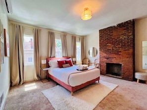 A bedroom with a large bed, a brick fireplace, and a view of the street through the window. - Mins to Stadiums - Spacious & Elegant (Pittsburgh)
