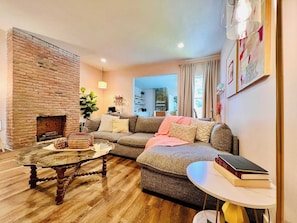 A cozy living room with a brick fireplace, a sectional sofa, a glass coffee table, and a small round table with books. - Mins to Stadiums - Spacious & Elegant (Pittsburgh)