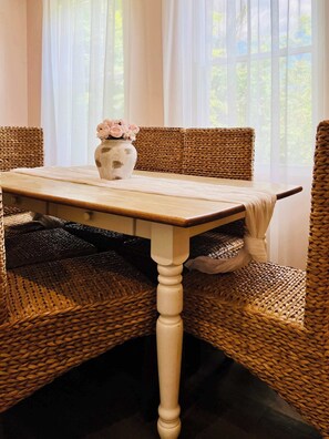 Un coin repas avec une table en bois et des chaises en osier, un vase rempli de fleurs et des rideaux légers.
