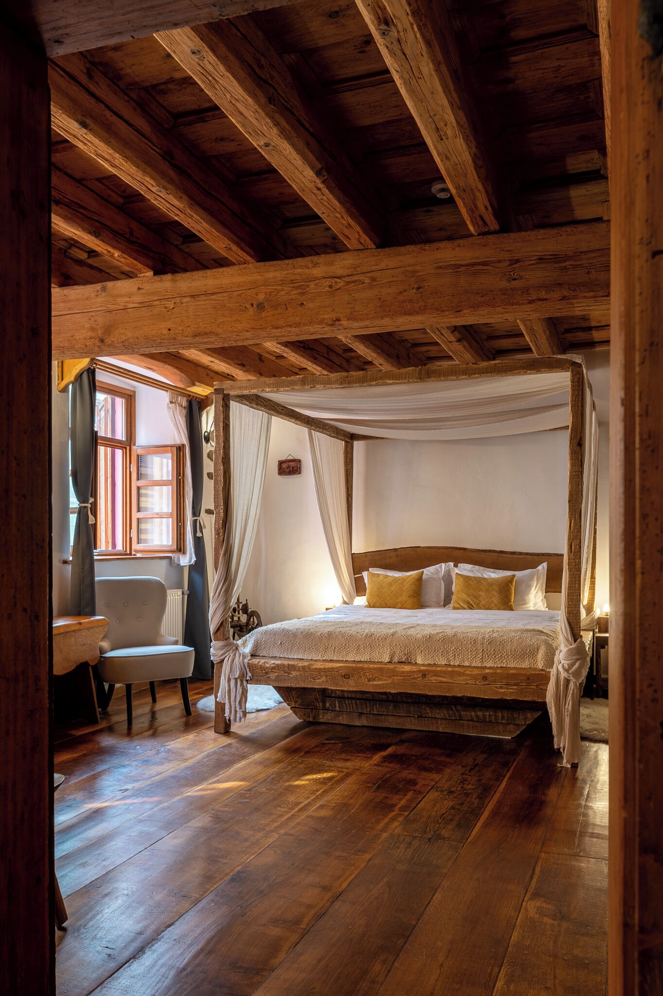 Ein Bett mit Baldachin und Holzkopf, ein kleiner Tisch und ein Stuhl in einem rustikalen Zimmer mit Holzbalken und einem Fenster.