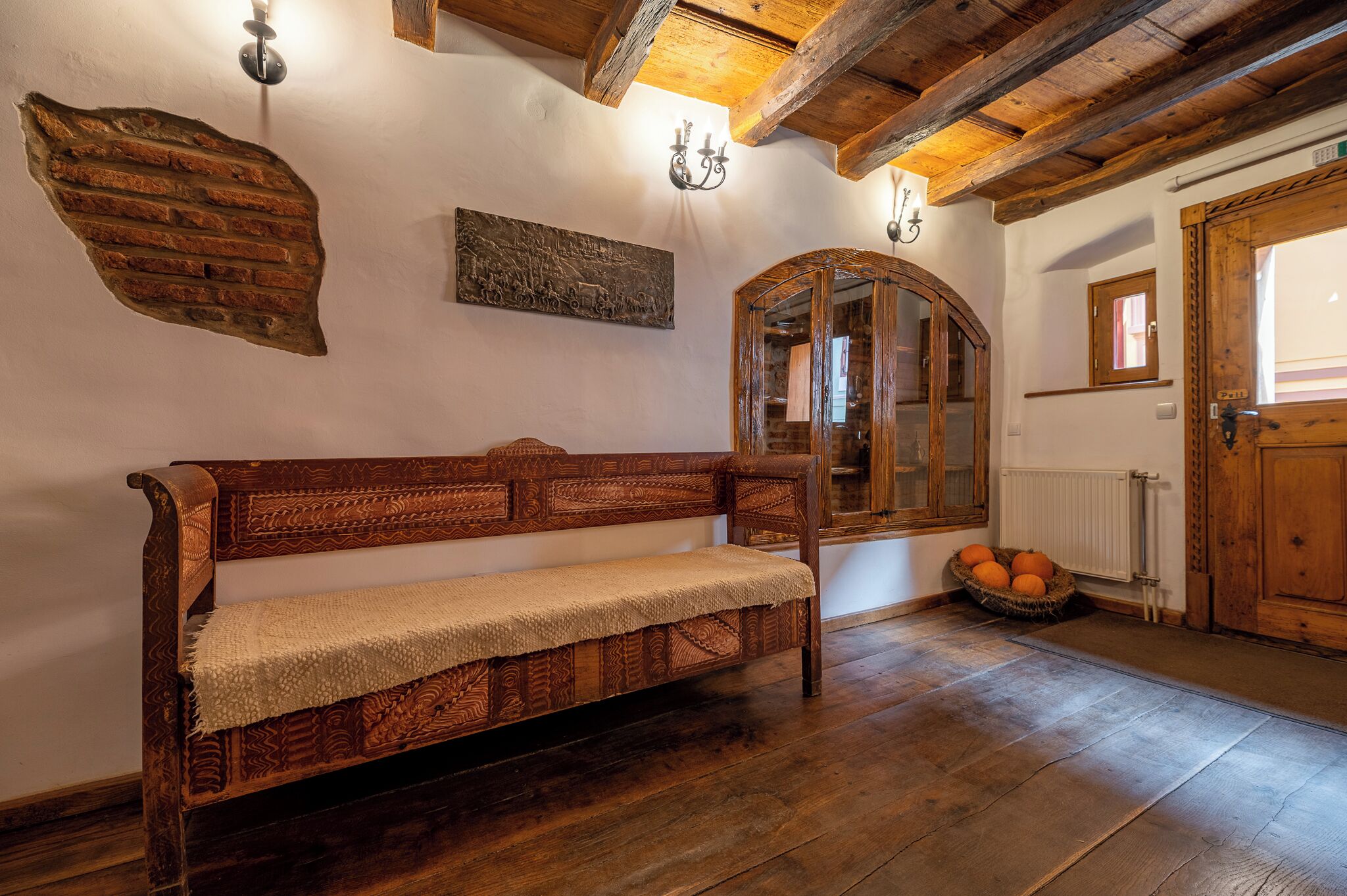 A wooden bench with a woven cover, a rustic wall with a brick-like texture, a wooden-framed mirror, and a basket of oranges on the floor.