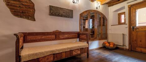 A wooden bench with a woven cover, a rustic wall with a brick-like texture, a wooden-framed mirror, and a basket of oranges on the floor.