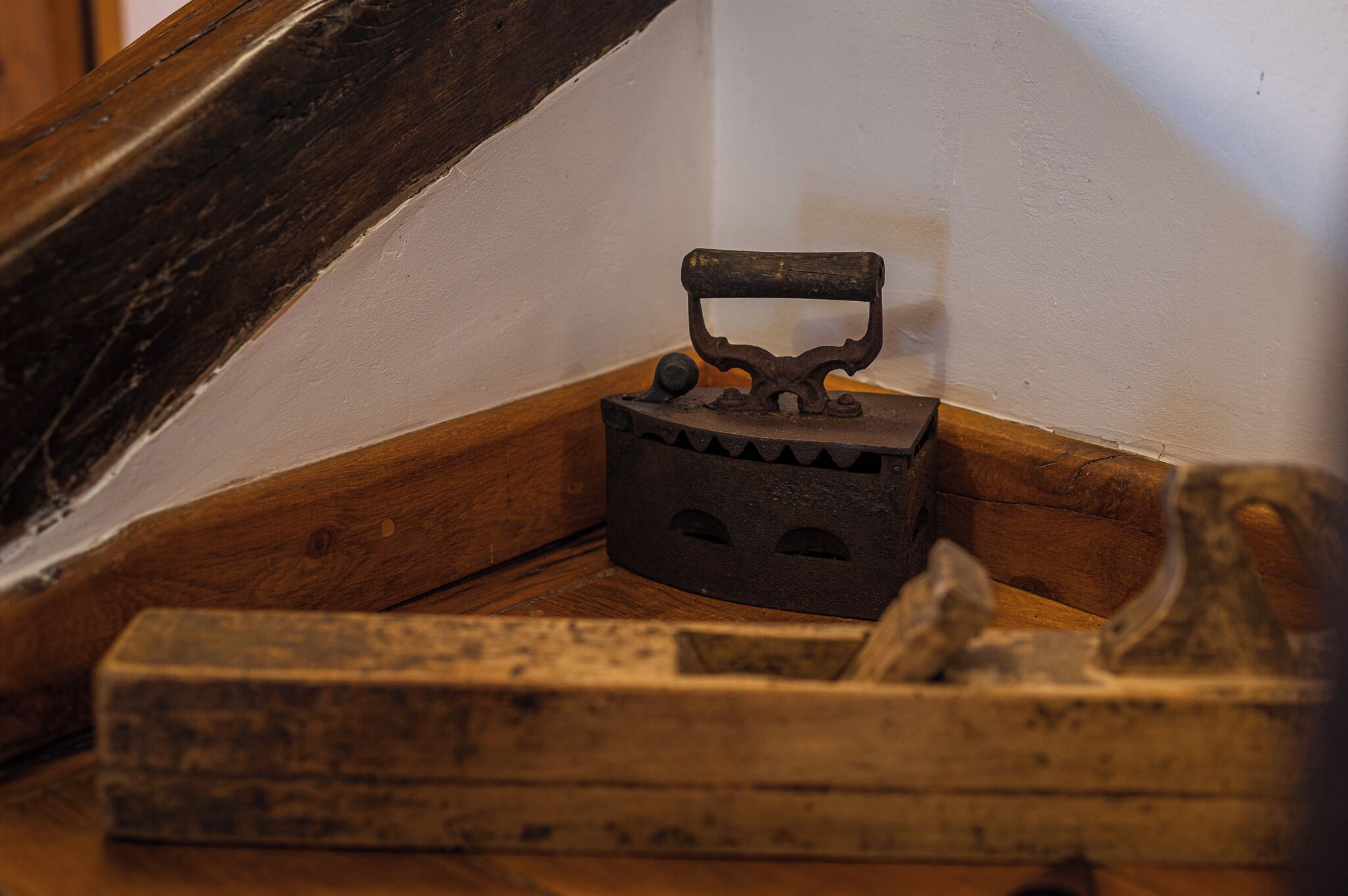 An antique iron with a wooden handle, placed on a wooden floor against a white wall.