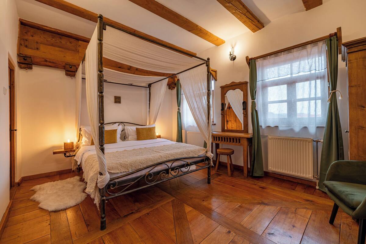 A four-poster bed with a canopy, wooden flooring, a small stool, a mirror, and a green chair in a room with wooden beams and a window with curtains.