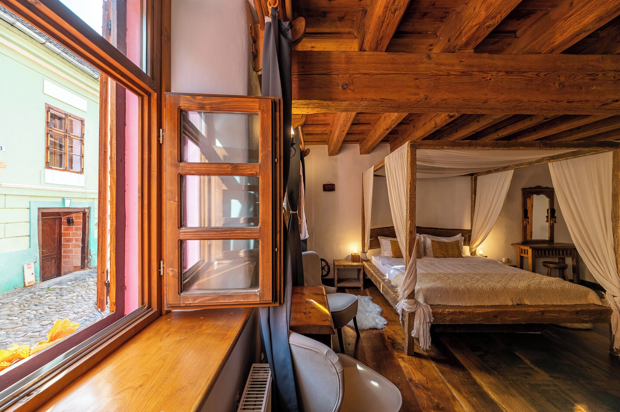 Una habitación acogedora con techo de madera, una cama con cortinas blancas, una mesita y un ventanal con vista a la calle.